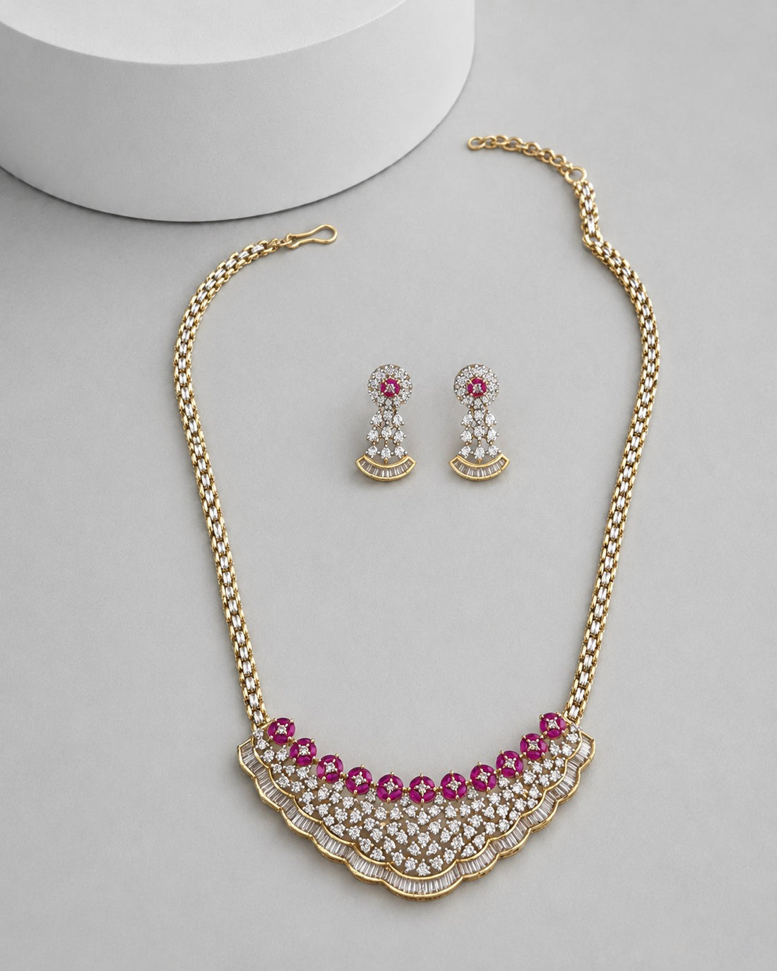 Gold-tone statement necklace and matching drop earrings with pink gemstone accents and clear rhinestone detailing, arranged on a light gray surface with a white cylindrical prop.