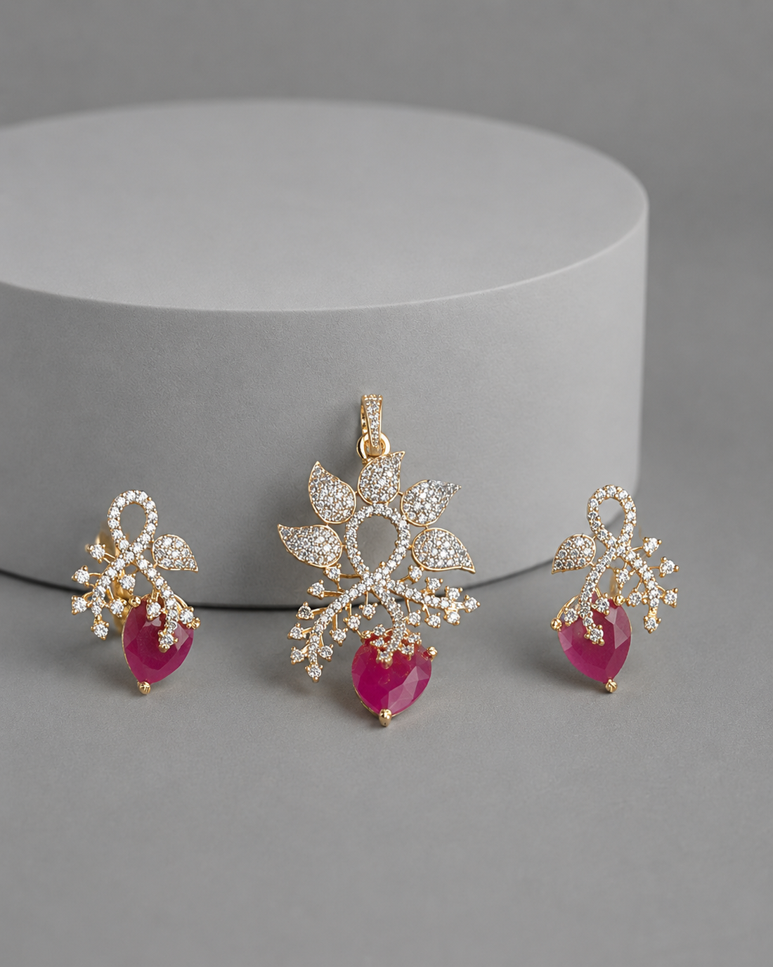 Gold-toned jewelry set featuring a central pendant and two matching drop earrings, decorated with clear pavé stones and finished with heart-shaped pink gemstone drops, displayed on a gray surface in front of a white cylindrical pedestal.
