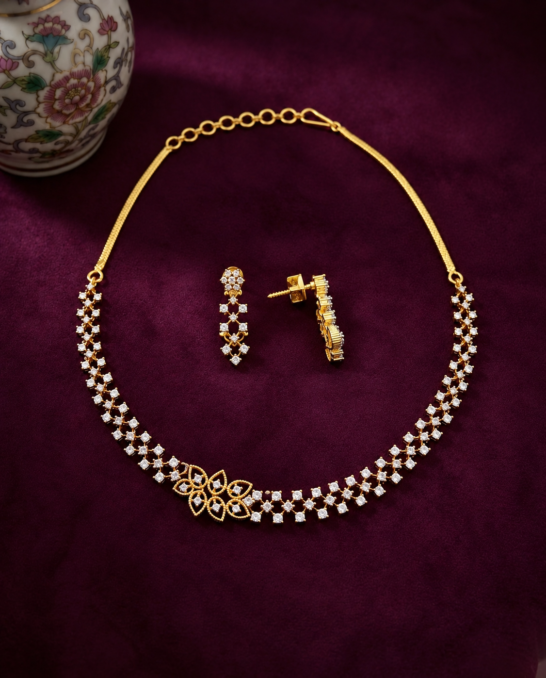 Gold-toned jewelry set featuring a double-row necklace with clear stones and a matching pair of drop earrings, displayed on a deep purple velvet surface with a decorative ceramic vessel partially visible in the corner.