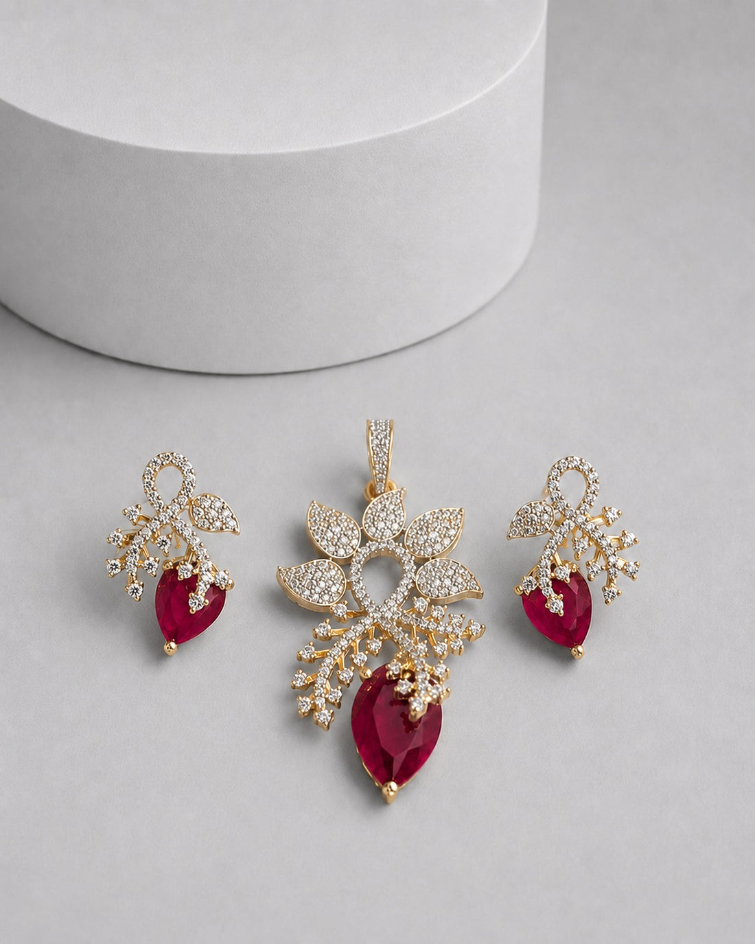 Gold-toned jewelry set featuring a pendant and two matching drop earrings, decorated with clear pavé stones and accented with deep red teardrop gemstones, displayed on a light gray surface with a white cylindrical prop.