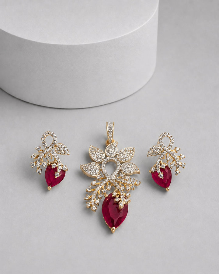 Gold-toned jewelry set featuring a pendant and two matching drop earrings, decorated with clear pavé stones and accented with deep red teardrop gemstones, displayed on a light gray surface with a white cylindrical prop.