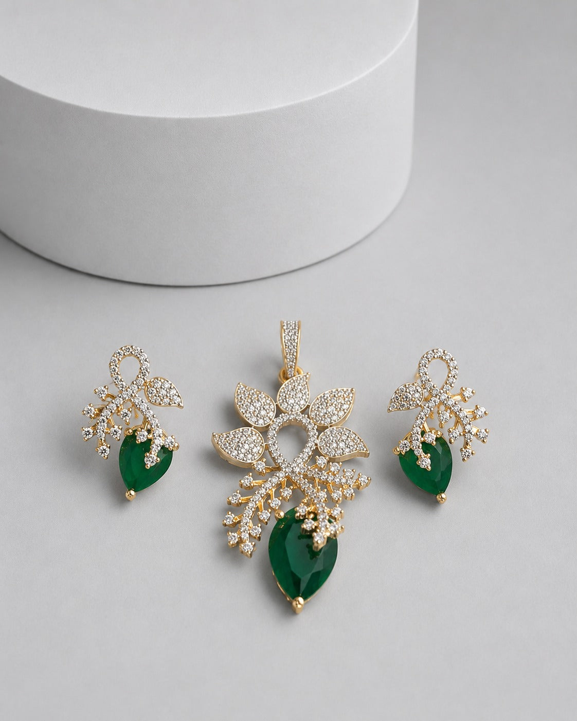 Gold-plated jewelry set featuring a pendant and matching earrings with clear pavé stones and deep green teardrop gemstones, displayed on a light gray surface with a white cylindrical prop.
