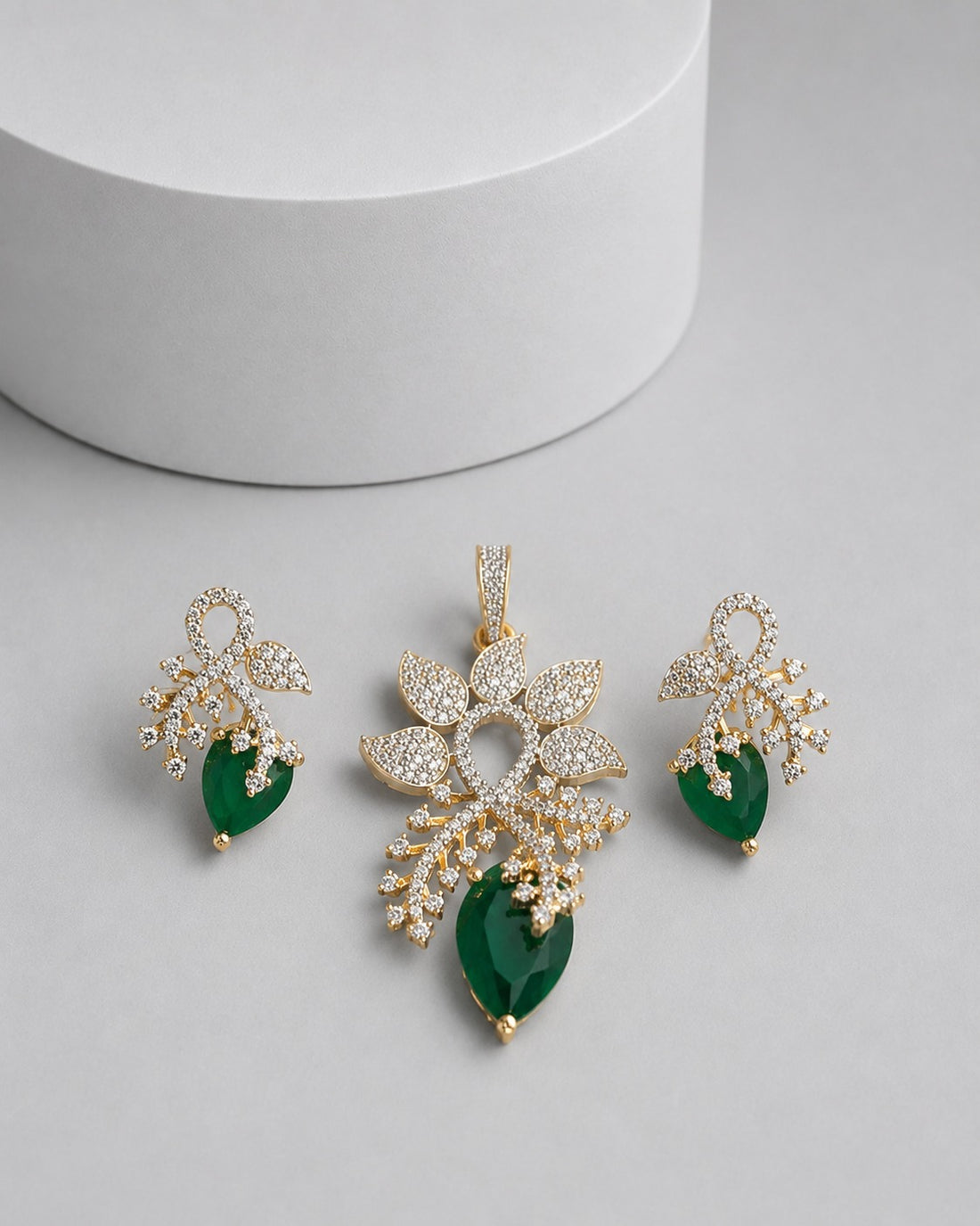 Gold-plated jewelry set featuring a pendant and matching earrings with clear pavé stones and deep green teardrop gemstones, displayed on a light gray surface with a white cylindrical prop.
