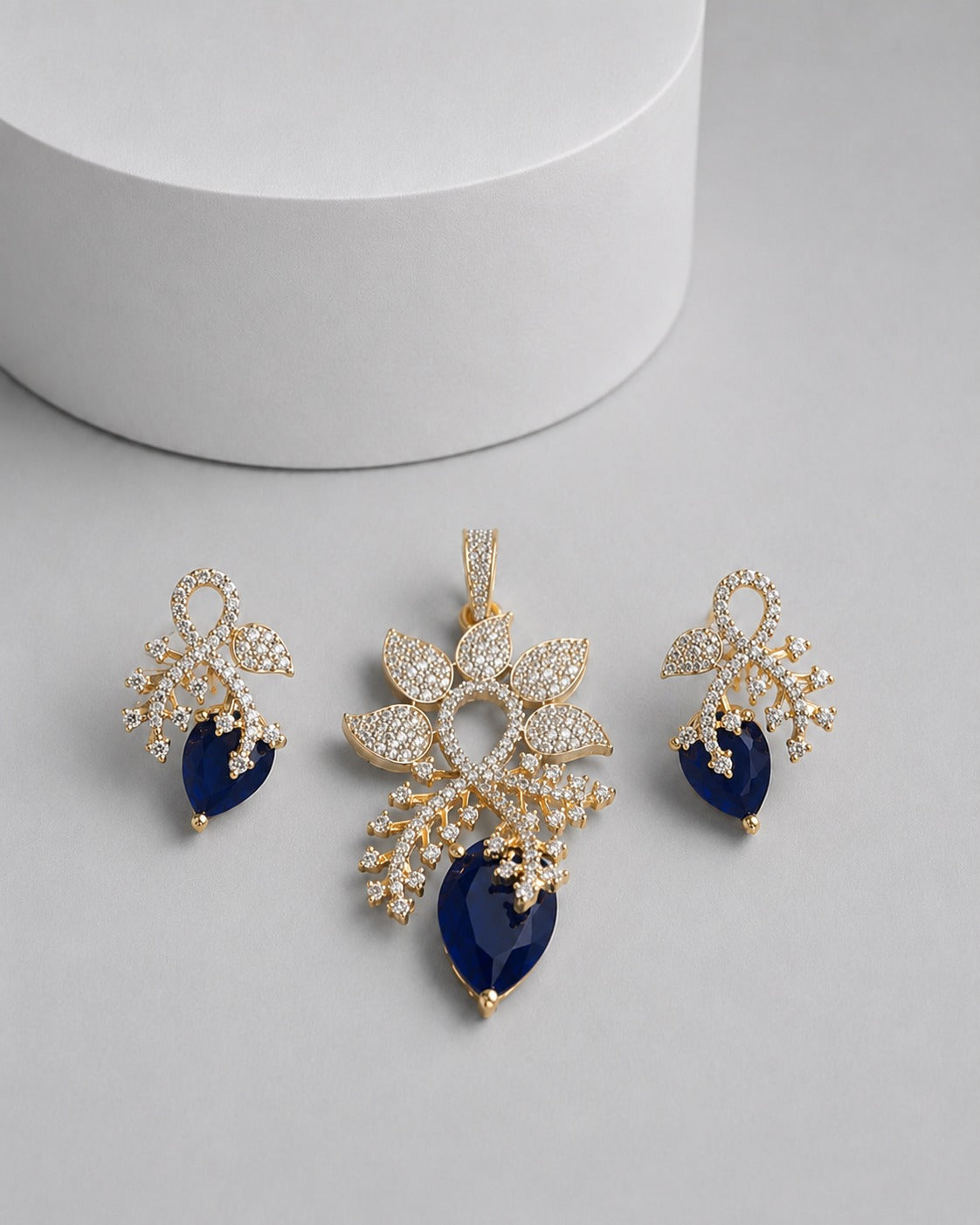 Gold-tone jewelry set featuring a floral pendant, matching earrings, and a small charm, embellished with clear pavé stones and accented with deep blue teardrop gemstones, displayed on a light gray surface with a white cylindrical pedestal.