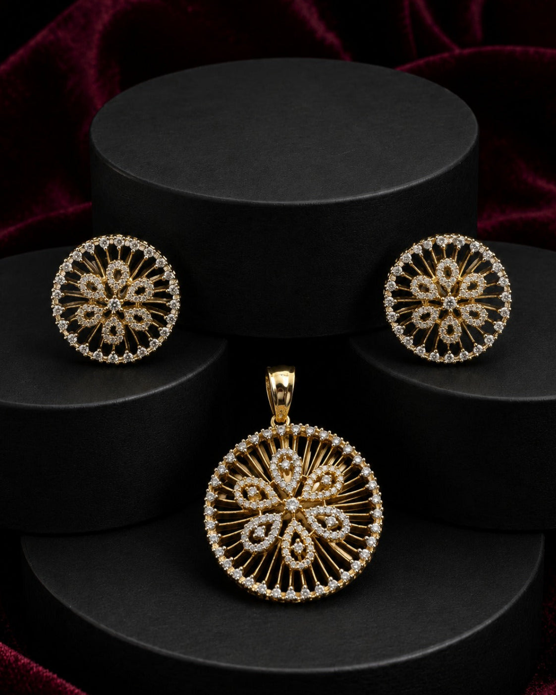 Gold-toned jewelry set featuring a round pendant and matching stud earrings with intricate filigree cutouts and small clear stones, displayed on black cylindrical pedestals against a dark red velvet background.