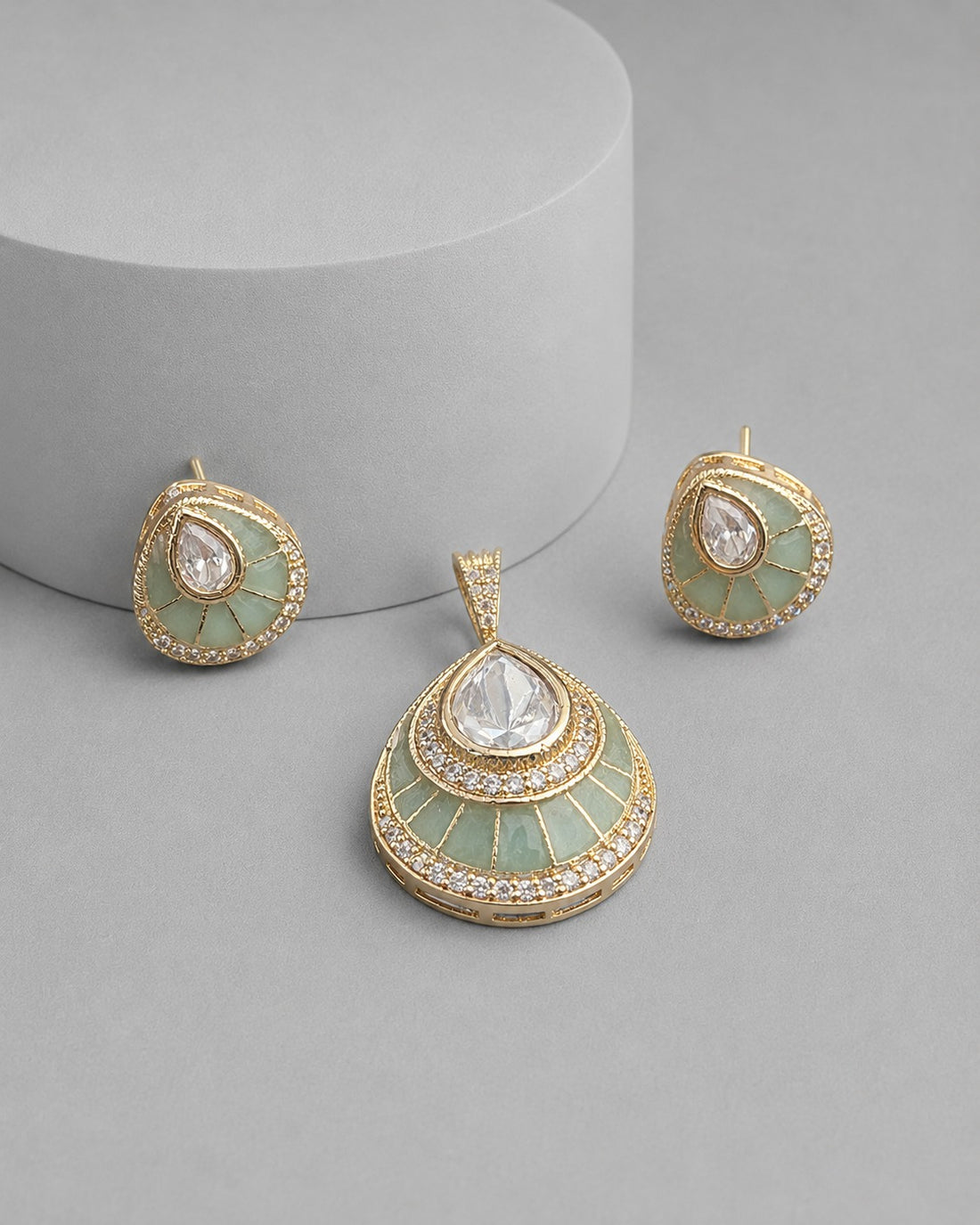 Gold-toned jewelry set featuring a teardrop-shaped pendant and matching stud earrings with pale mint green enamel, clear teardrop gemstones, and small clear stones around the edges, displayed on a light gray surface with a cylindrical pedestal.