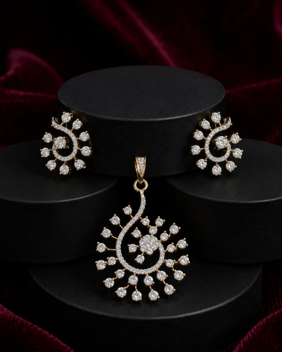 Gold-toned jewelry set featuring a teardrop pendant and matching stud earrings with pavé-set clear stones, displayed on black cylindrical pedestals against a dark red velvet background.