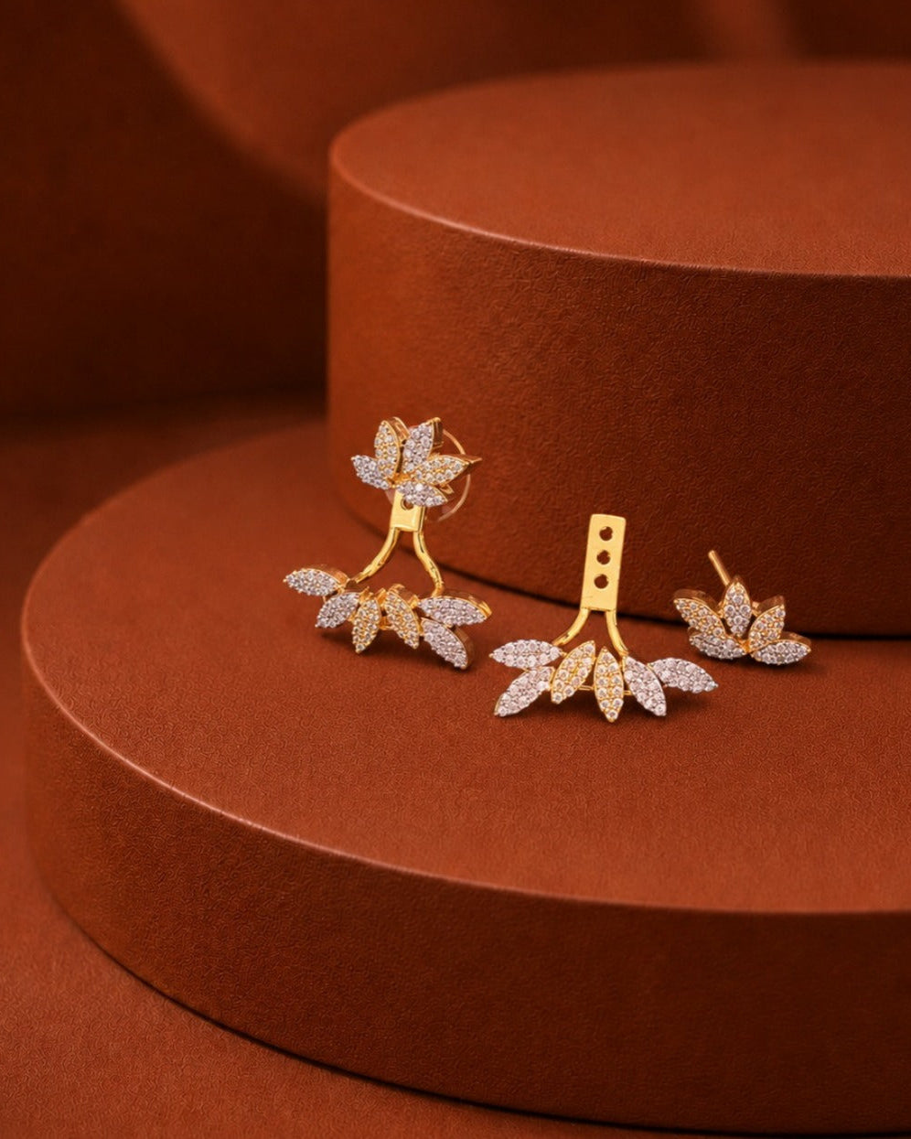 Gold-toned leaf-shaped earrings with pavé-set clear stones, arranged on a warm brown cylindrical display stand.