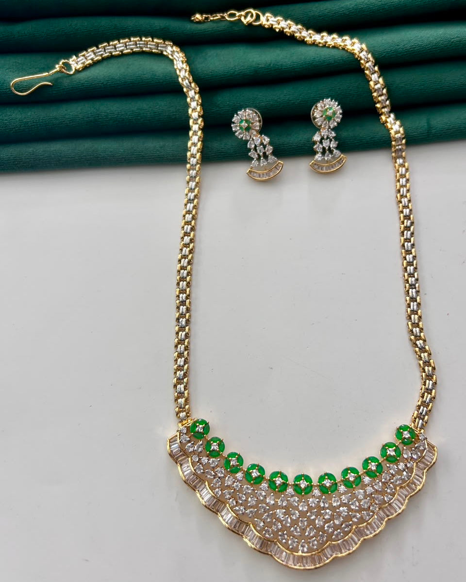 Gold-toned necklace and matching earrings set with green and clear rhinestones, displayed on a white surface with a dark green fabric backdrop.