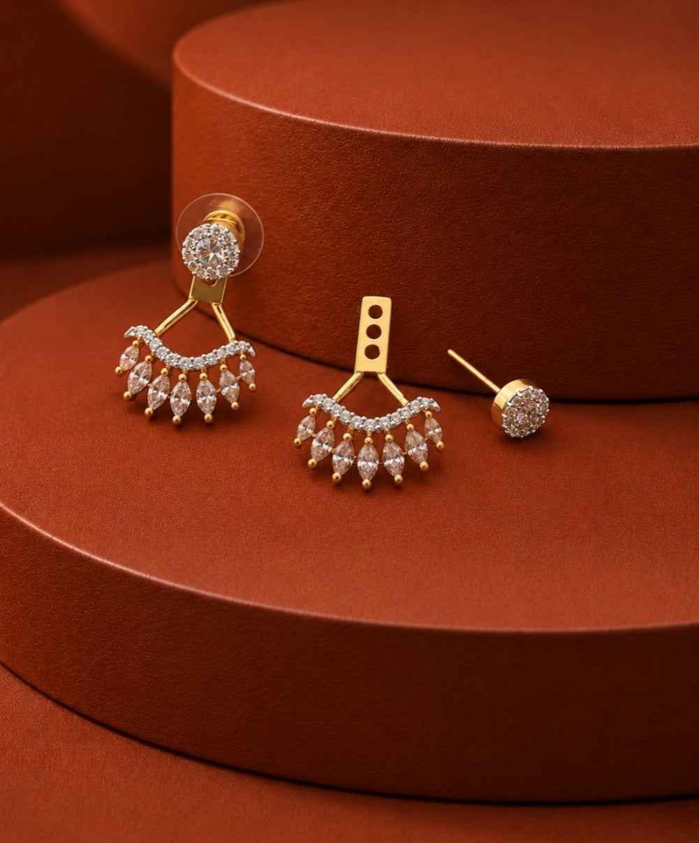 Gold-toned statement earrings with dangling crystal-like drops and a round stud variant, displayed on a warm brown cylindrical pedestal.