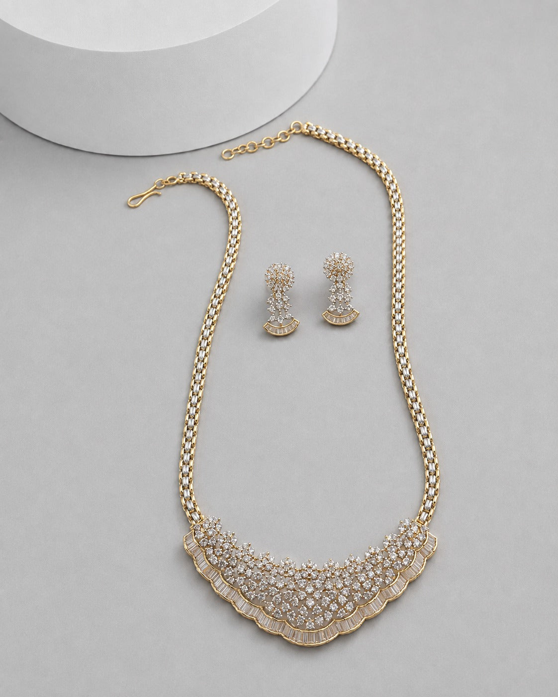 Gold-toned statement necklace and matching drop earrings with clear rhinestone embellishments, arranged on a light gray surface with a white cylindrical prop in the background.