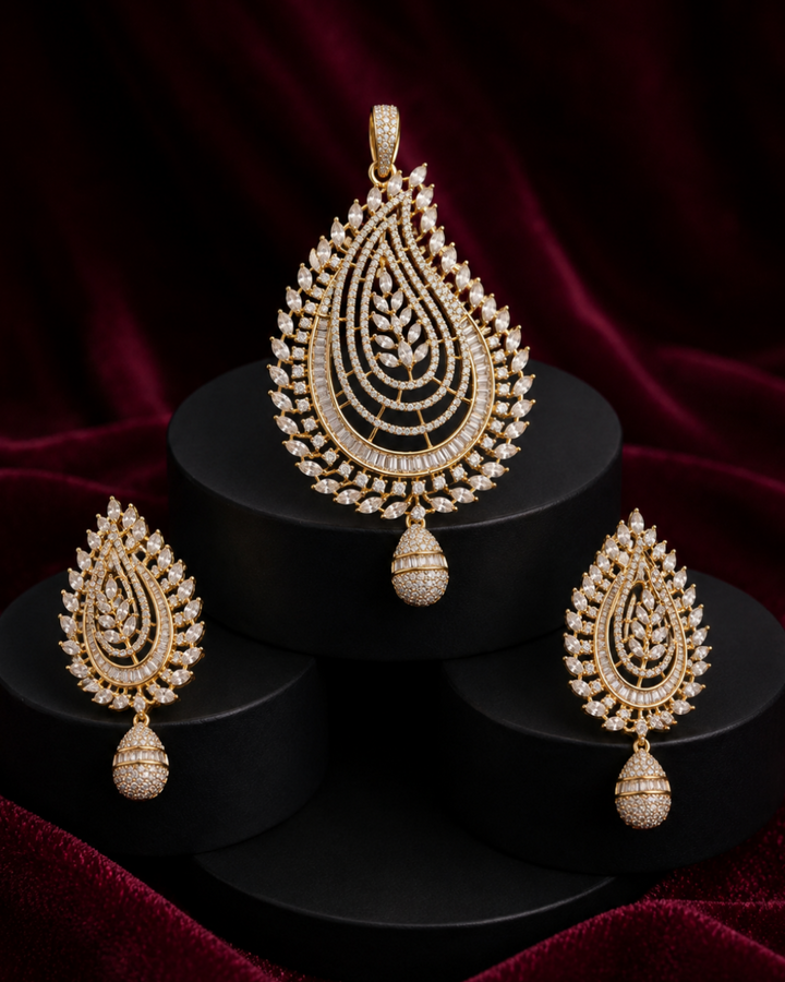 Gold-toned teardrop pendant necklace with matching drop earrings, displayed on black cylindrical pedestals against a deep burgundy velvet background.