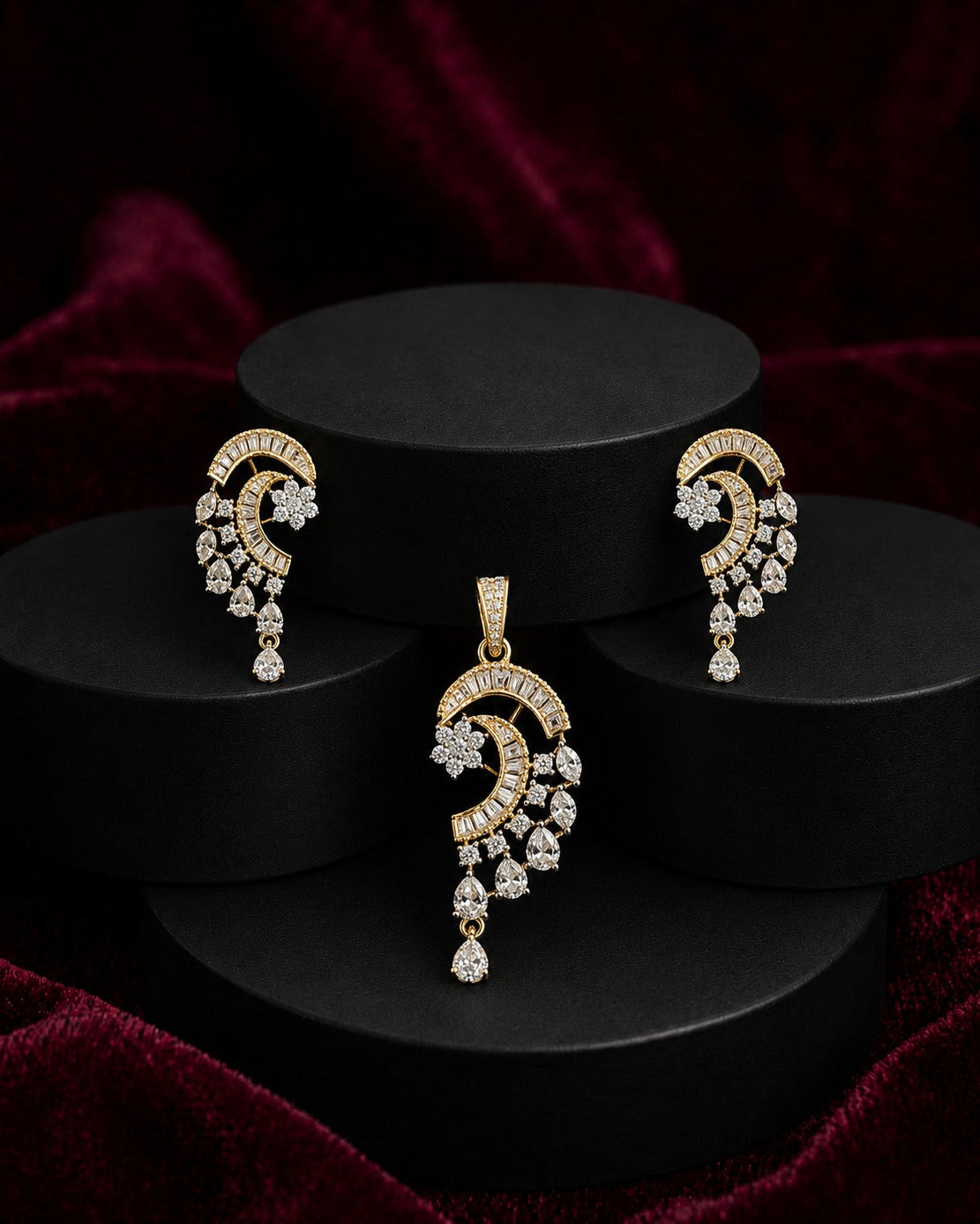 Gold-toned traditional Indian jewelry set featuring a crescent-shaped pendant and matching drop earrings, embellished with clear stones, displayed on black cylindrical pedestals against a dark red velvet background.