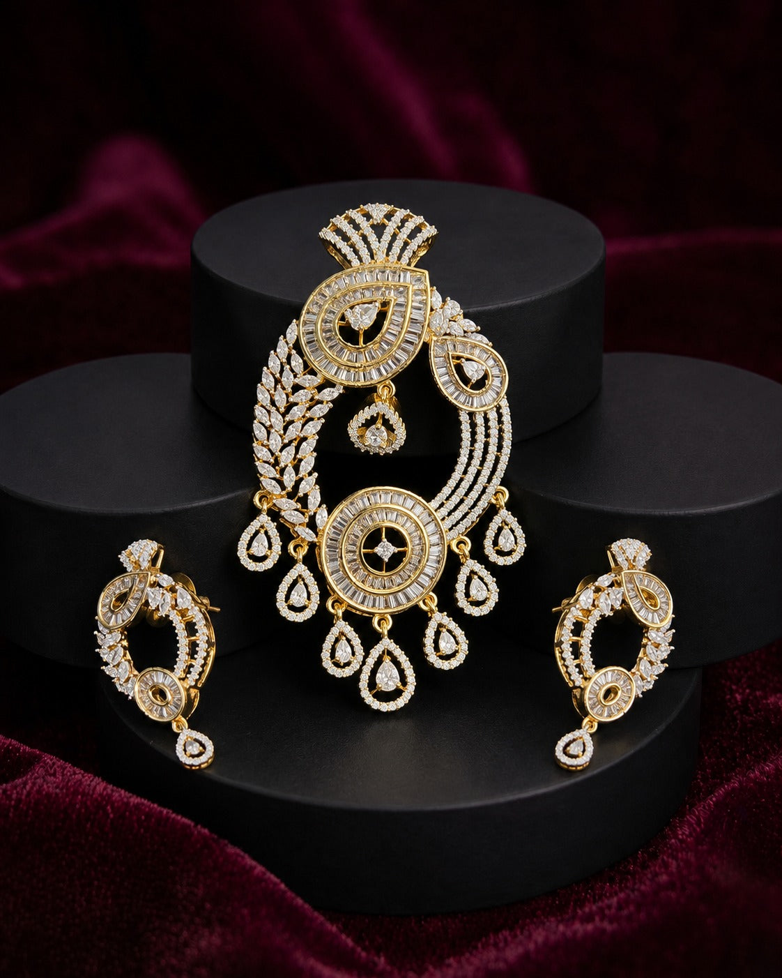 Gold-toned traditional Indian jewelry set featuring a large ornate pendant, matching earrings, and a small necklace, all embellished with clear stones, displayed on black cylindrical pedestals against a dark burgundy velvet background.