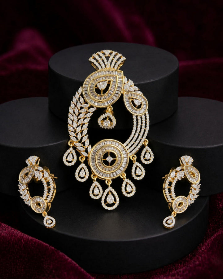 Gold-toned traditional Indian jewelry set featuring a large ornate pendant, matching earrings, and a small necklace, all embellished with clear stones, displayed on black cylindrical pedestals against a dark burgundy velvet background.