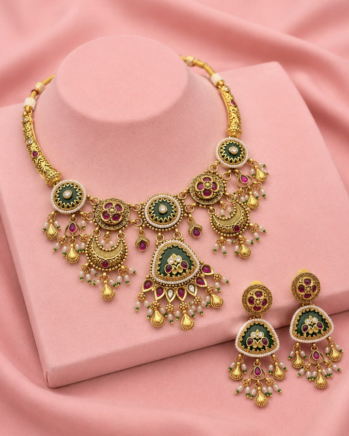 Gold-toned traditional Indian jewelry set featuring a statement necklace and matching earrings with green enamel, red stone accents, and pearl-like white beads, displayed on a pink mannequin stand and pink fabric background.