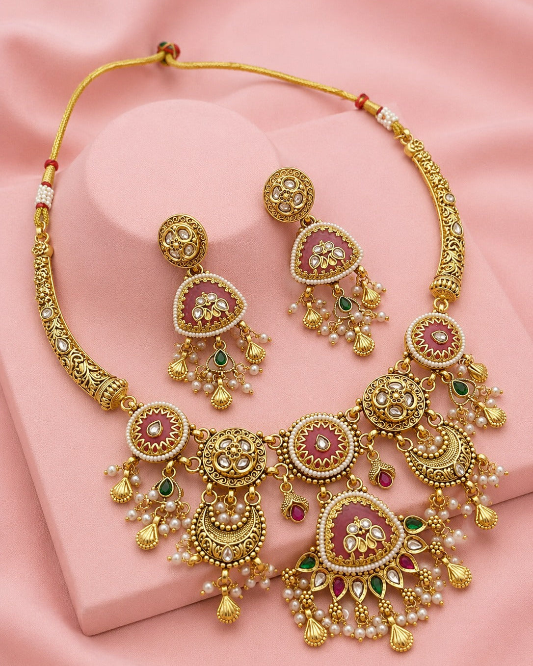 Gold-toned traditional jewelry set featuring a necklace and matching earrings with pink stone centers, white pearl-like beads, and small green and red accent stones, displayed on a pink fabric background.