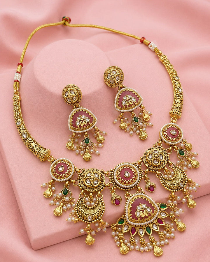 Gold-toned traditional jewelry set featuring a necklace and matching earrings with pink stone centers, white pearl-like beads, and small green and red accent stones, displayed on a pink fabric background.