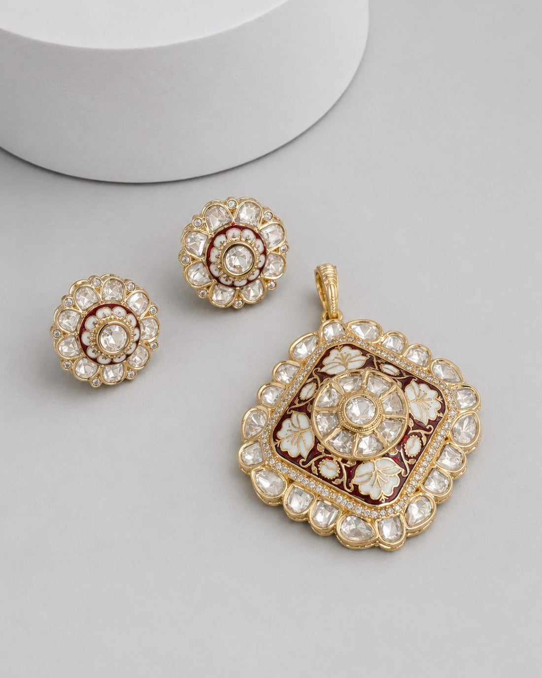 Gold-toned traditional jewelry set featuring a square pendant and two matching round earrings, decorated with clear stones and dark red enamel on a light gray background.