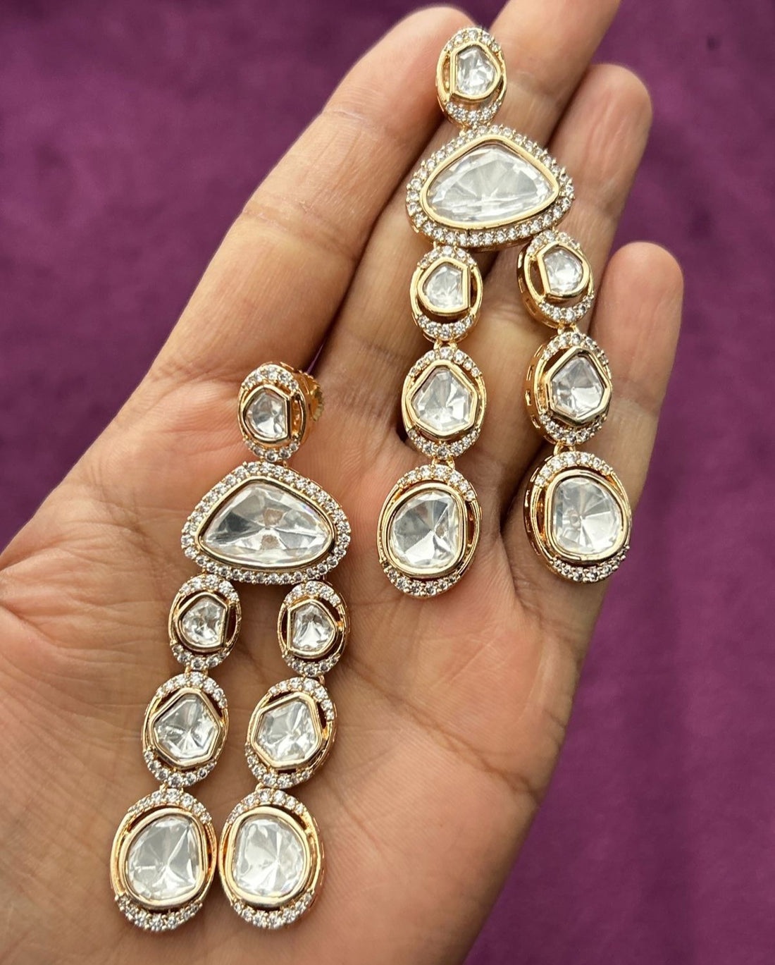 Gold and crystal earrings held in a hand against a purple background