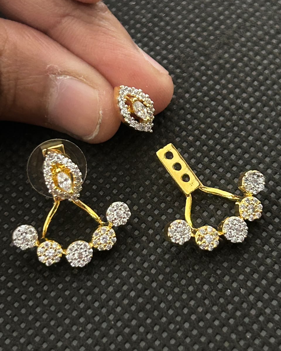 Gold and crystal pave stud earrings shown on a textured black background, with a person's fingers holding one earring for scale.
