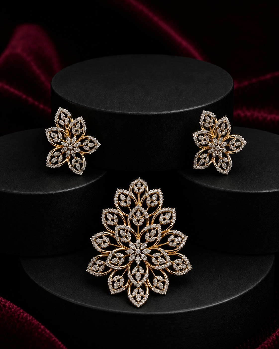 Gold and diamond jewelry set featuring a large floral brooch and matching stud earrings, displayed on black cylindrical pedestals against a dark red velvet background.