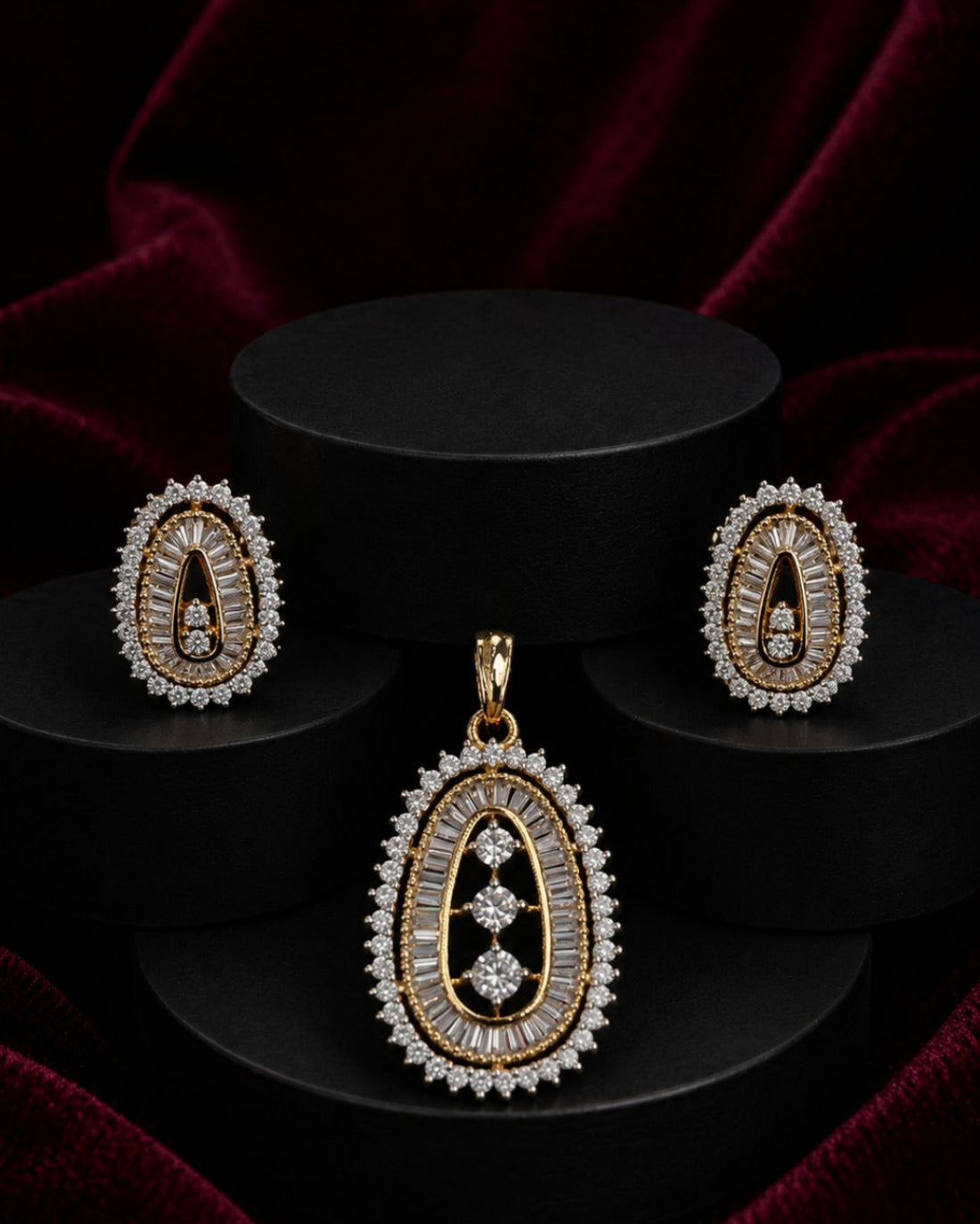 Gold and diamond jewelry set featuring a teardrop pendant and matching earrings, displayed on black cylindrical pedestals against a dark red velvet background.