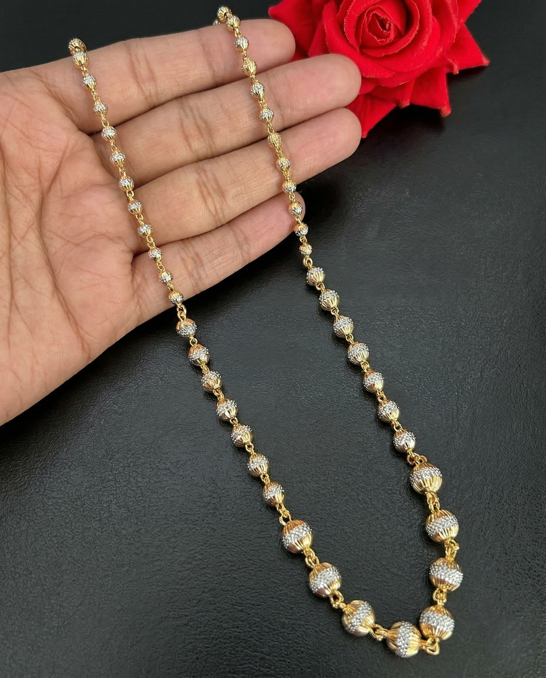 Gold and diamond necklace held by a hand with a red rose in the background on a dark surface