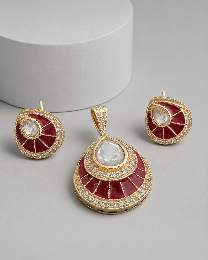Gold and red jewelry set featuring a teardrop pendant and two matching round earrings, decorated with clear stones and set against a neutral gray background with a white cylinder prop.