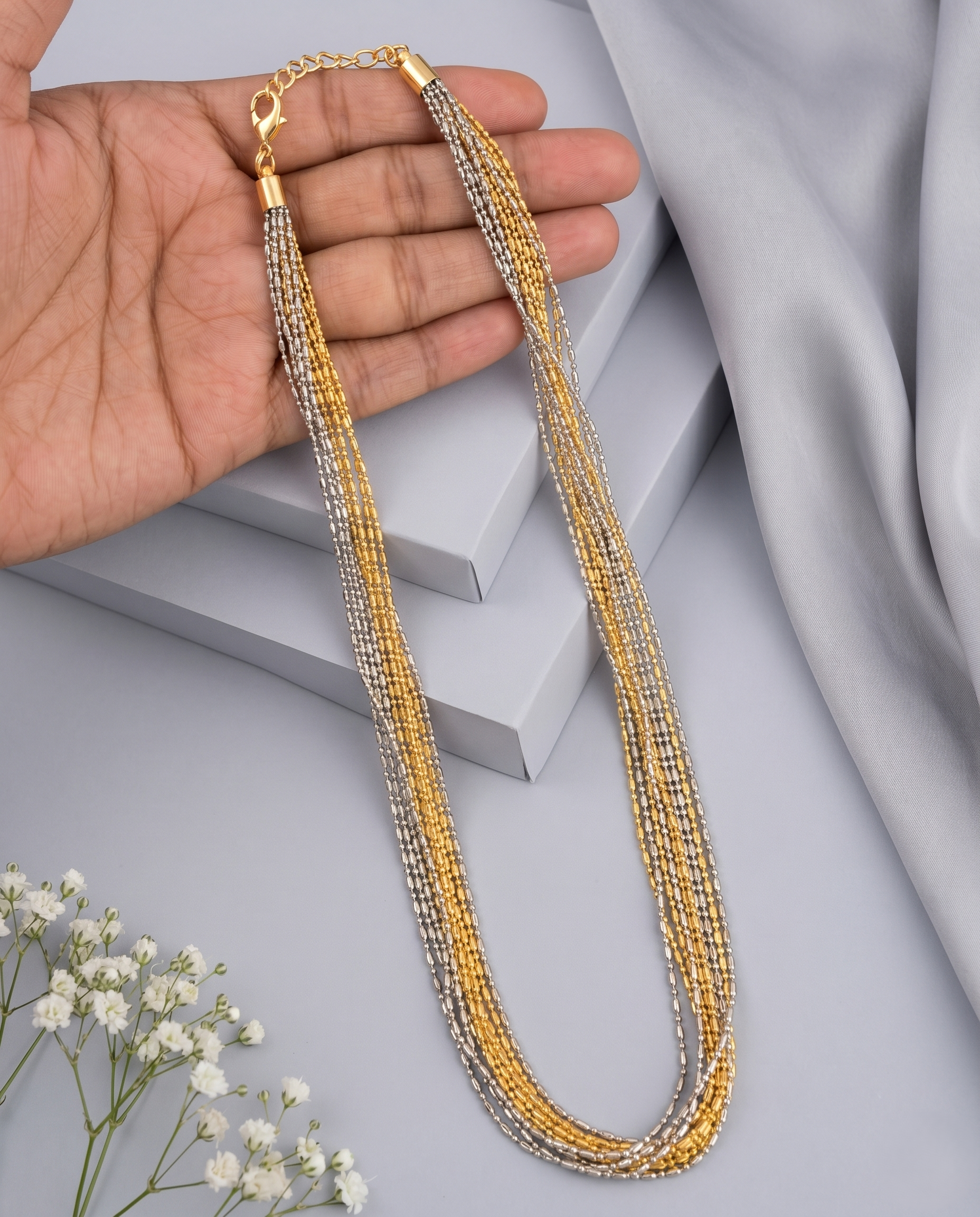 Gold and silver beaded necklace held by a hand on a light gray background with white flowers.