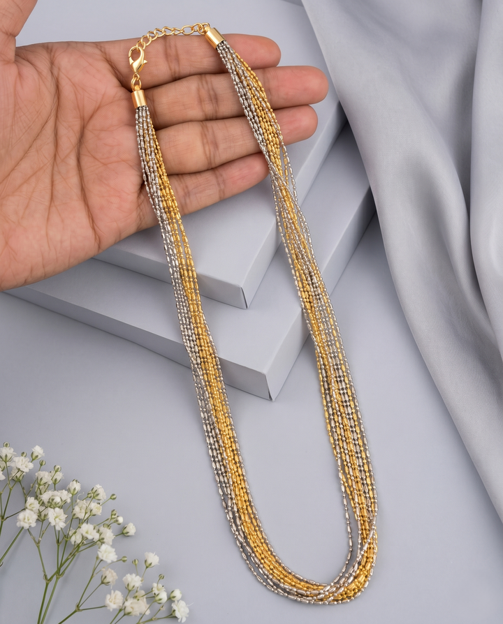 Gold and silver beaded necklace held by a hand on a light gray background with white flowers.