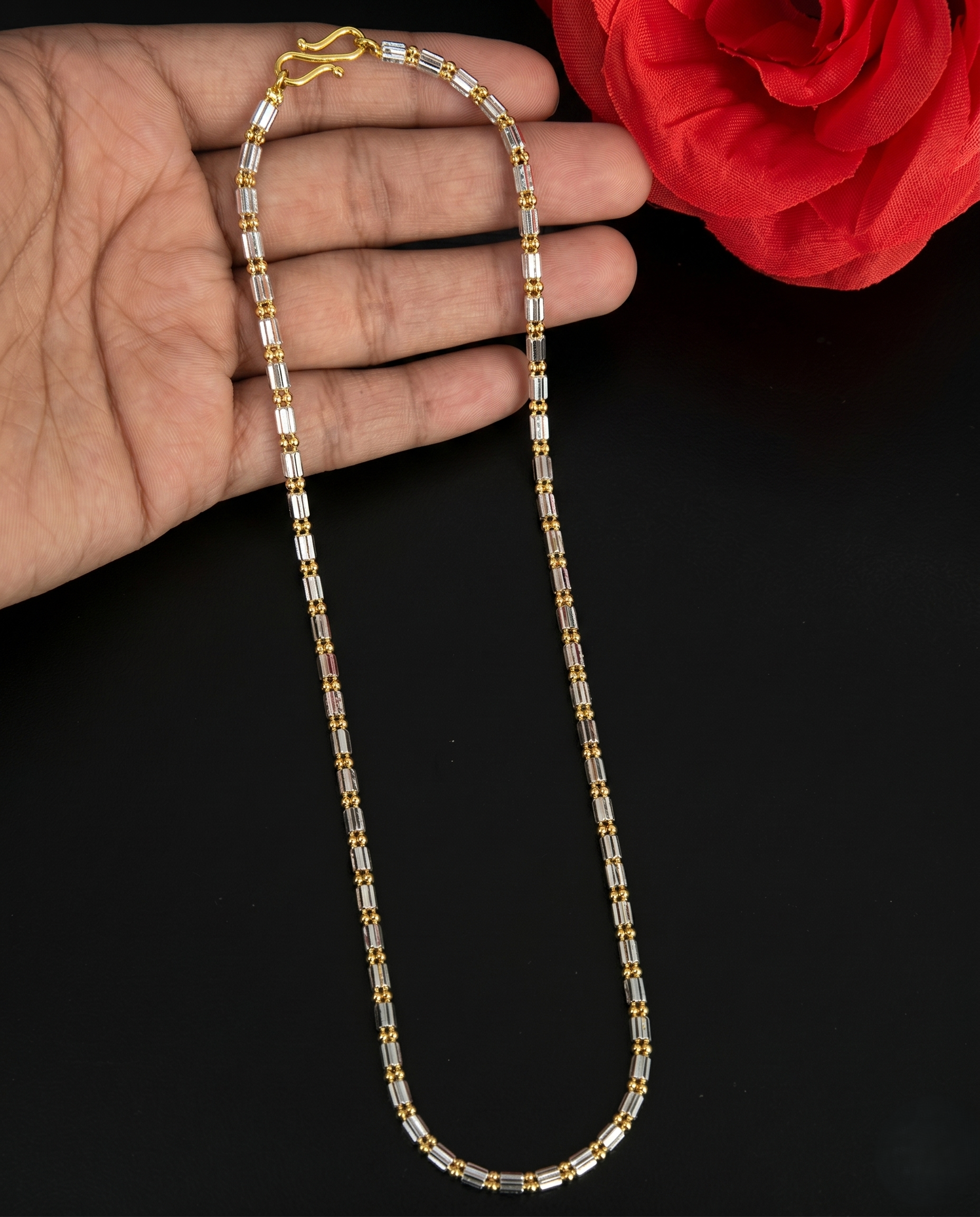 Gold and silver beaded necklace held by a hand with a red flower in the background