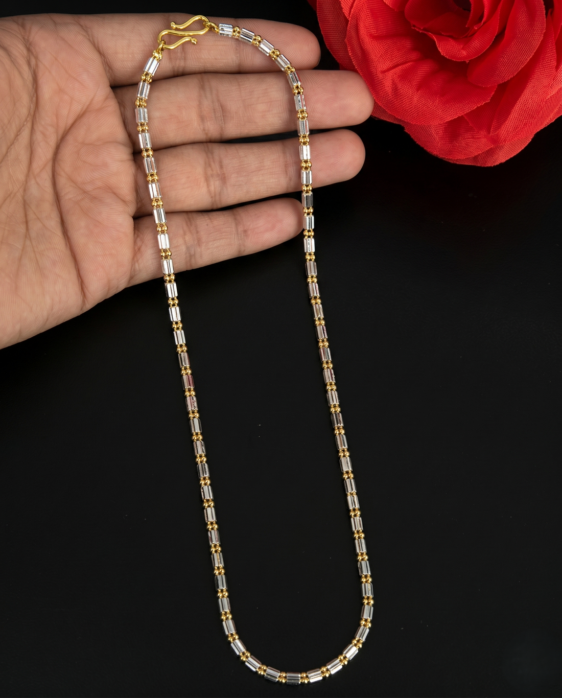 Gold and silver beaded necklace held by a hand with a red flower in the background