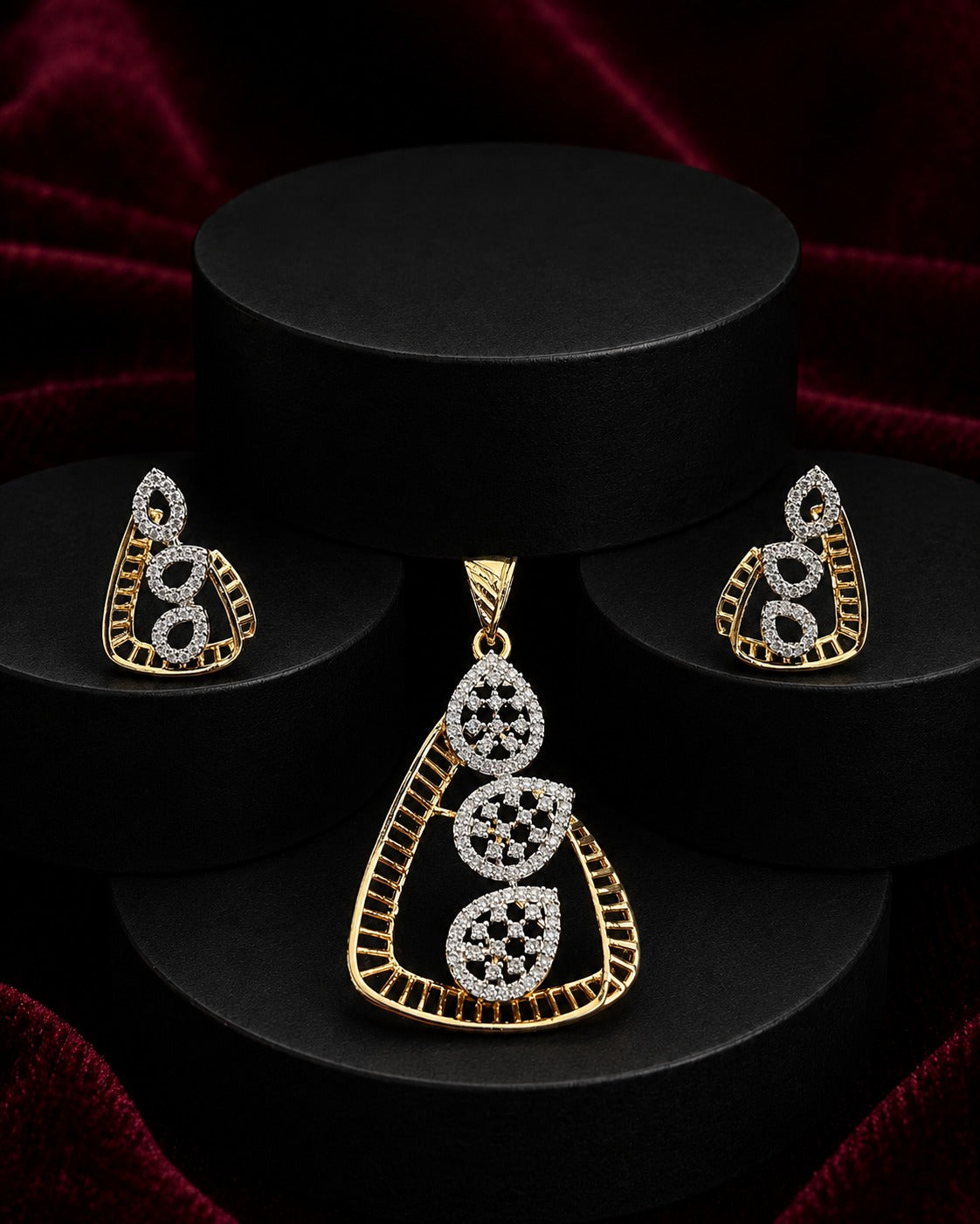 Gold and white stone jewelry set featuring a teardrop pendant and matching earrings, displayed on black cylindrical pedestals against a dark red velvet background.