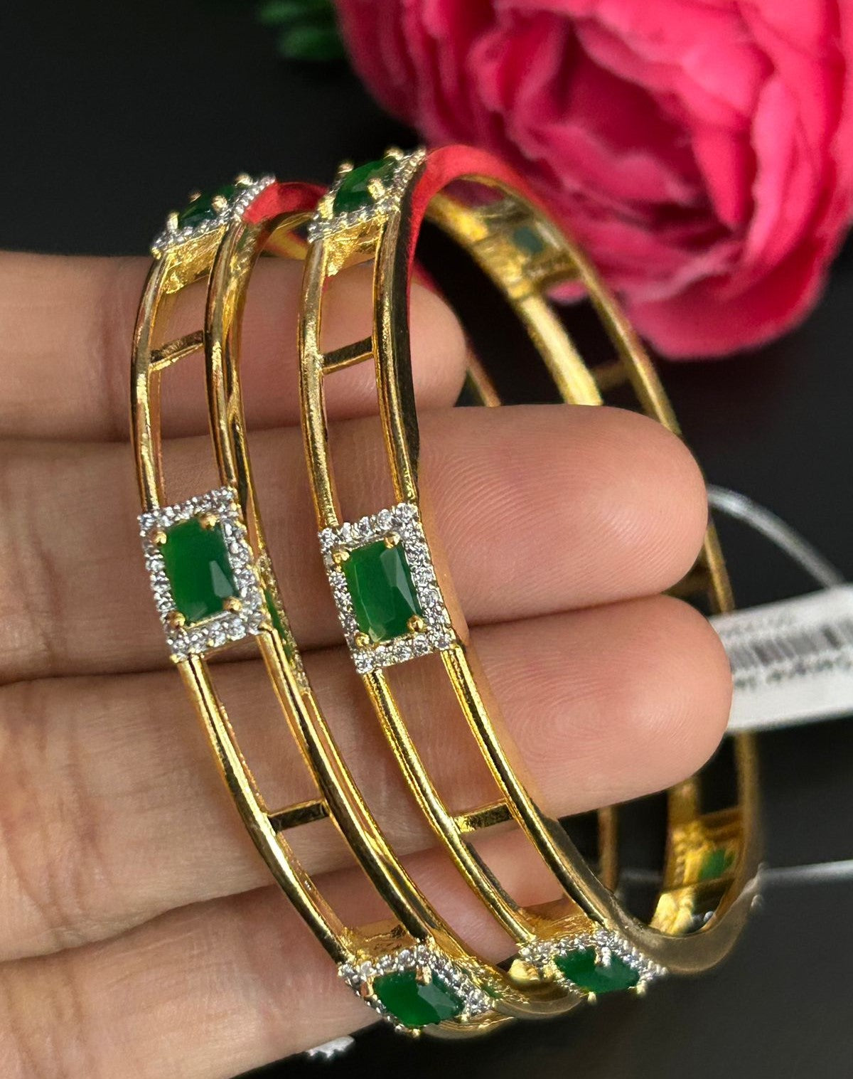 Gold bangles with green gemstones held by a hand, with a pink flower in the background.