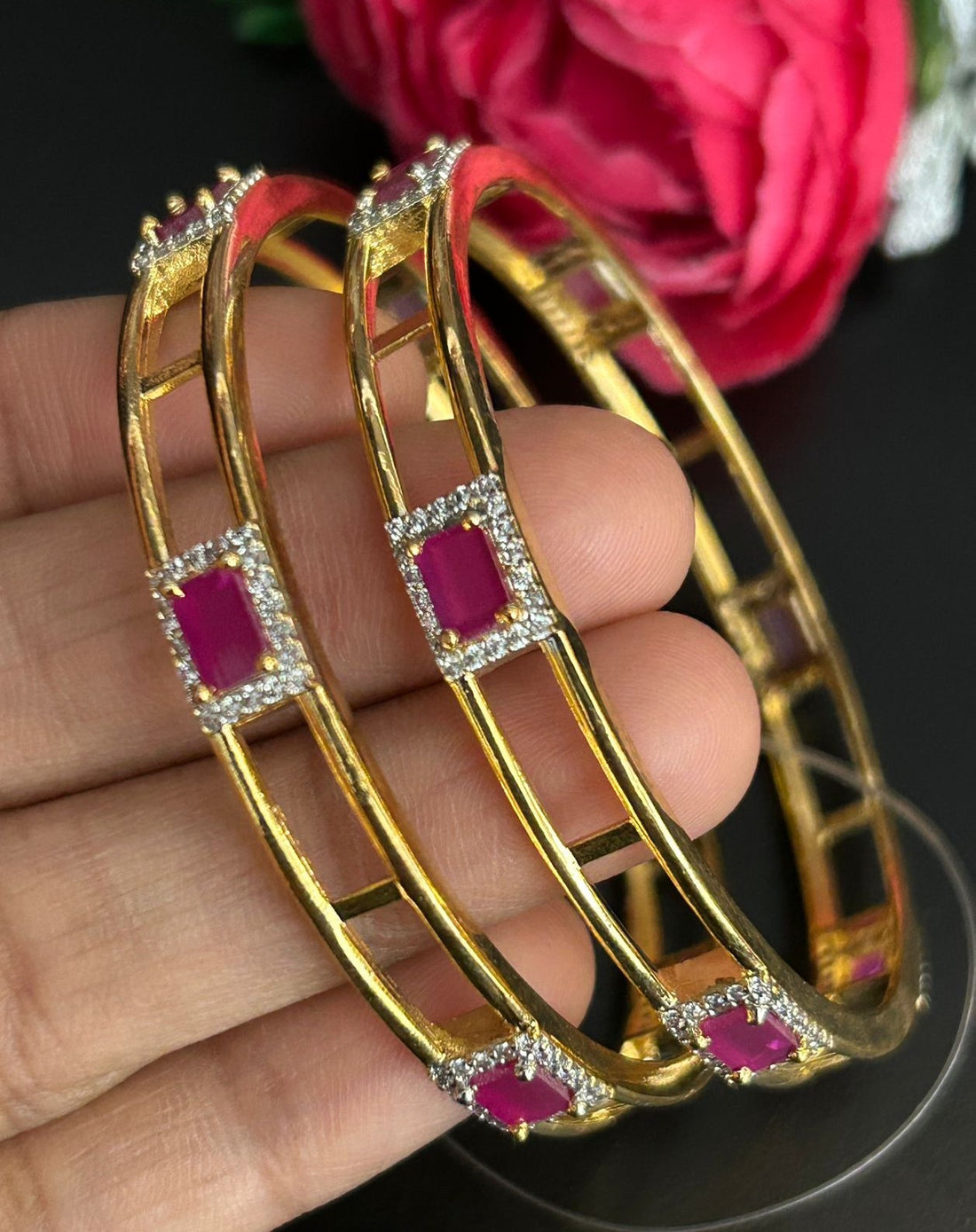 Gold bangles with red stones on a hand, against a dark background with pink flowers.