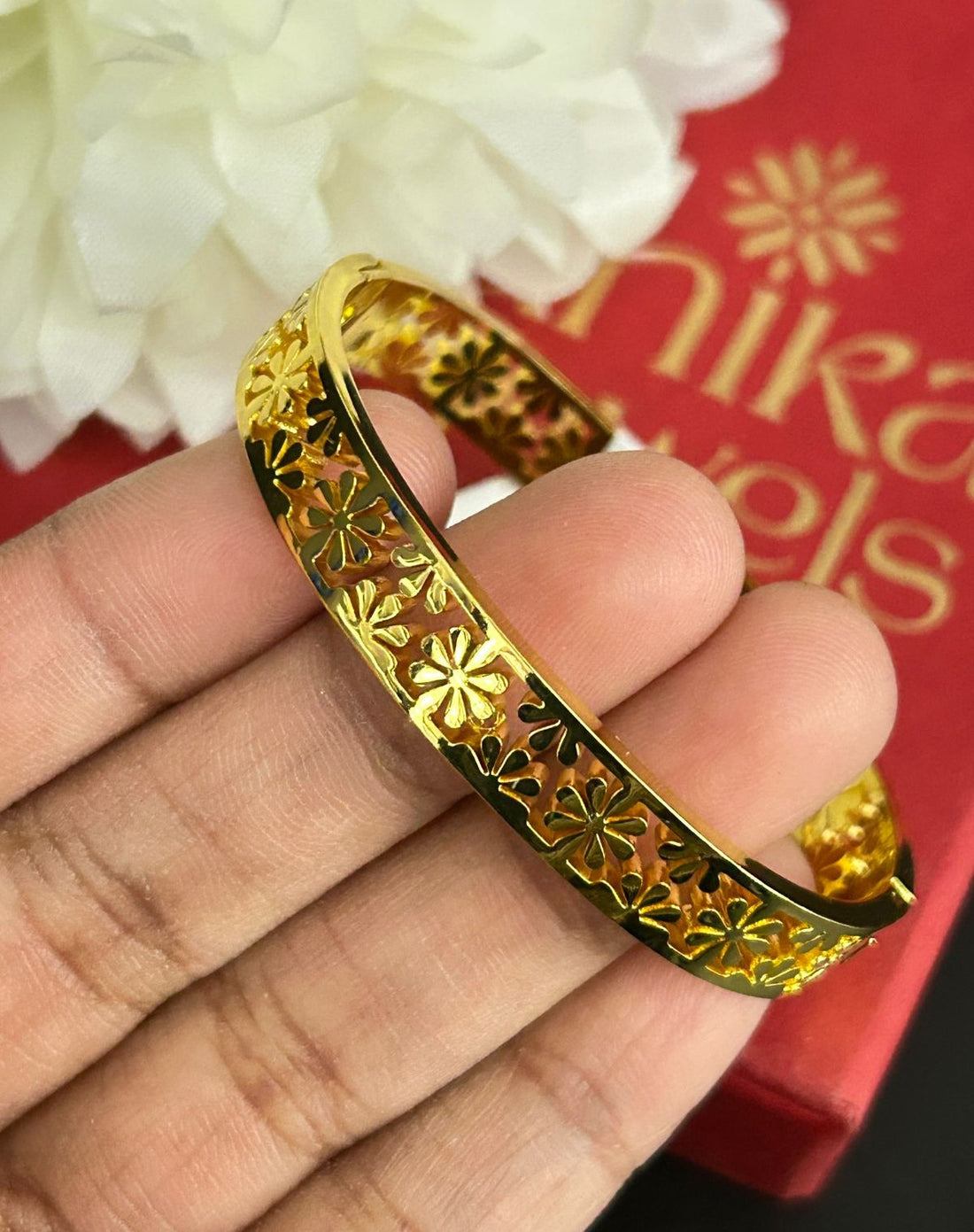 Gold bracelet held by a hand with a red box and white flowers in the background