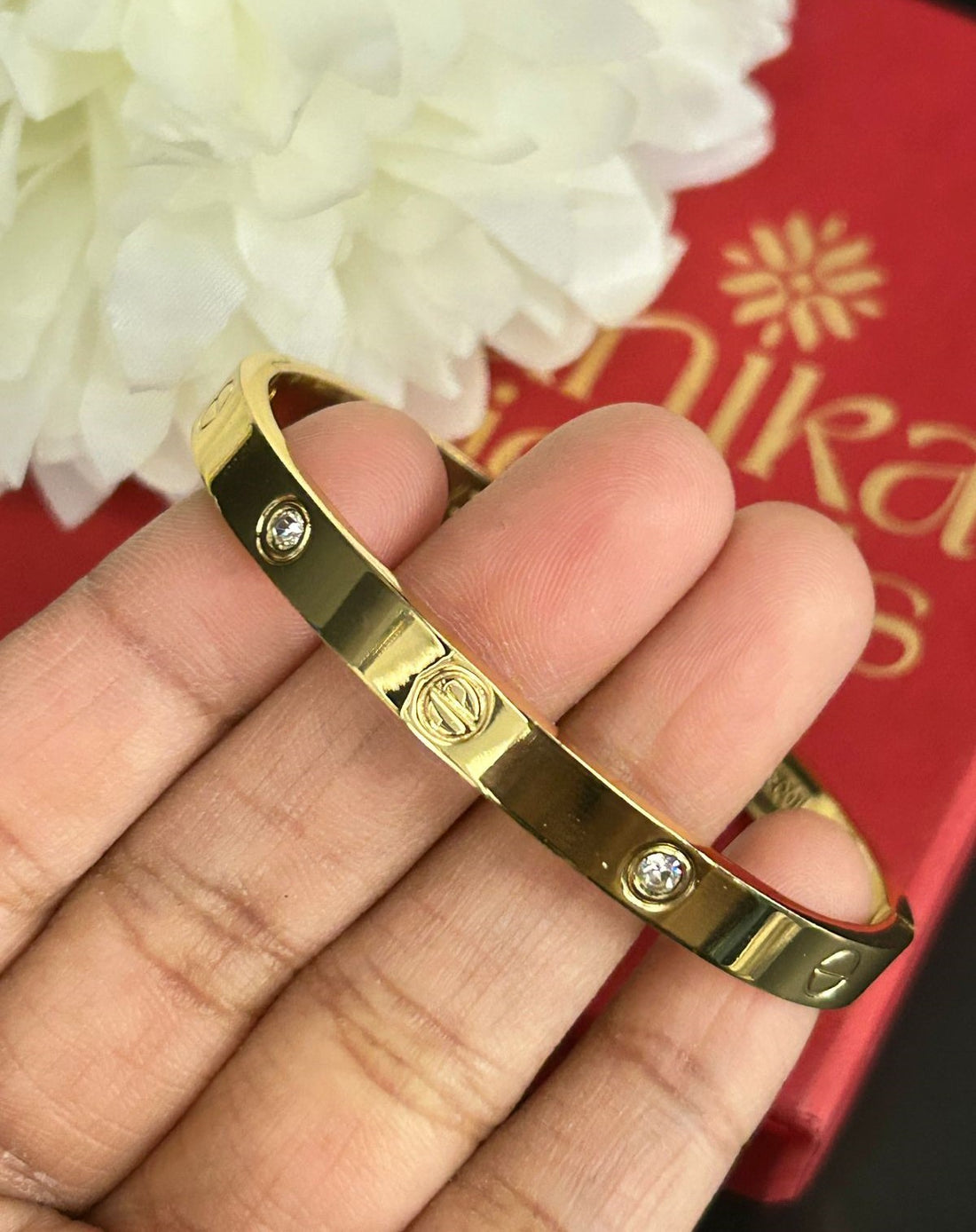 Gold bracelet with diamond accents on a hand, against a red background with 'NIDHIKA JEWELS' branding.