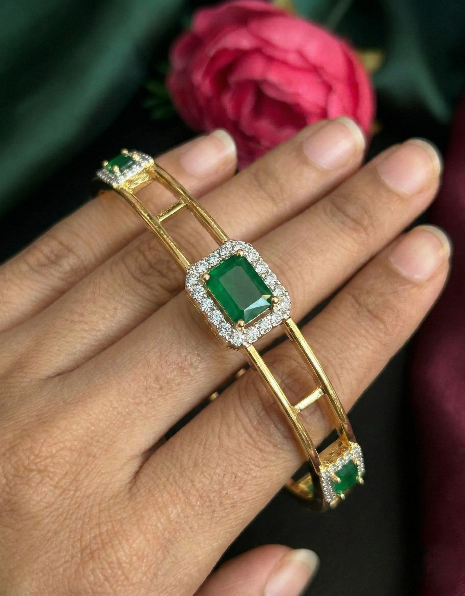 Gold bracelet with emerald stones on a hand, with a pink flower in the background