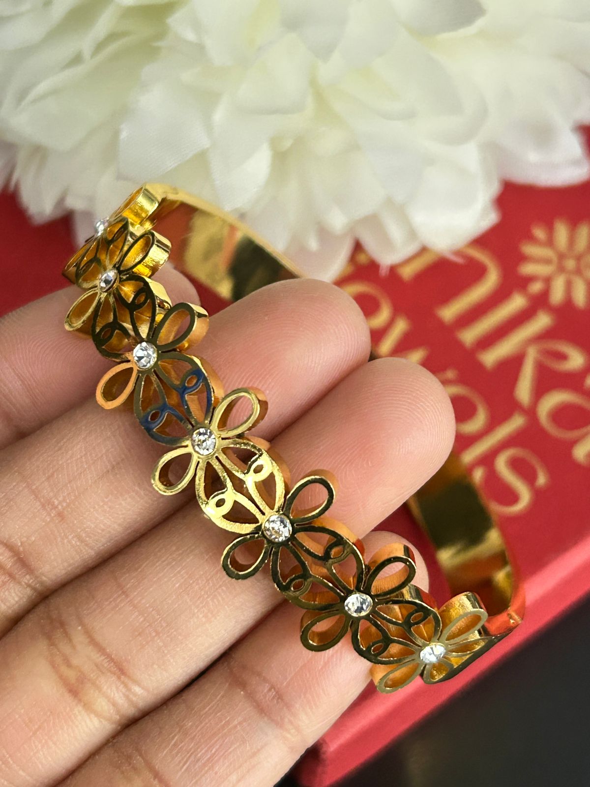 Gold bracelet with floral designs held by a hand, against a red background with floral elements.
