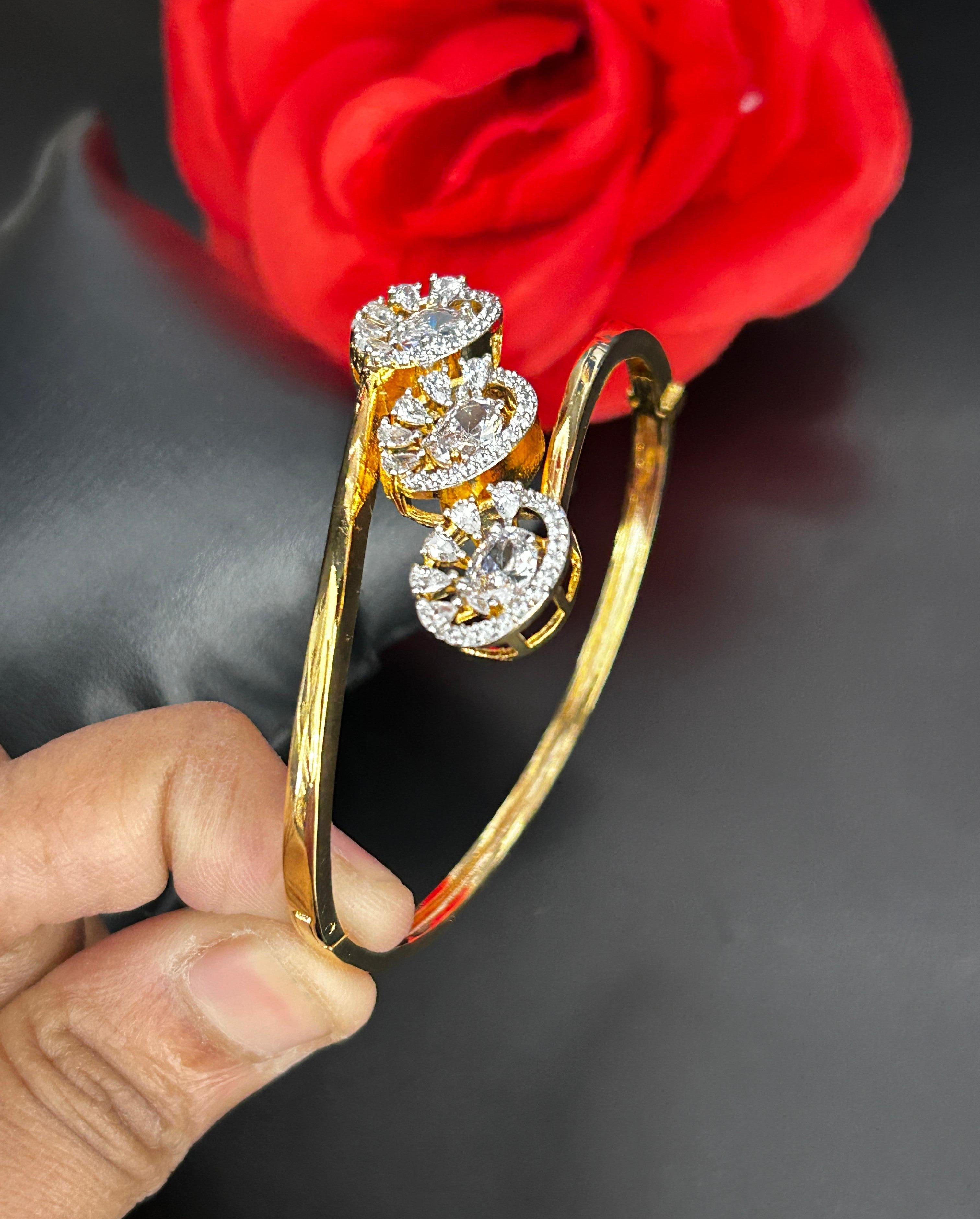 Gold bracelet with three diamond-like stones held by a hand against a red flower on black fabric background