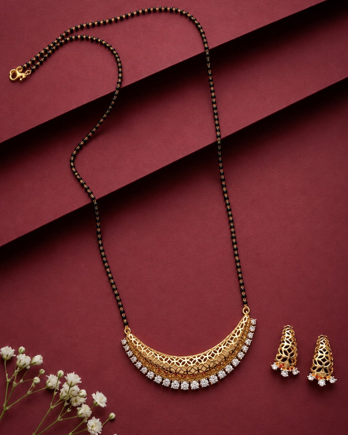Gold crescent-shaped mangalsutra with a black beaded chain and white stones, displayed with matching earrings on a dark red background with small white flowers.