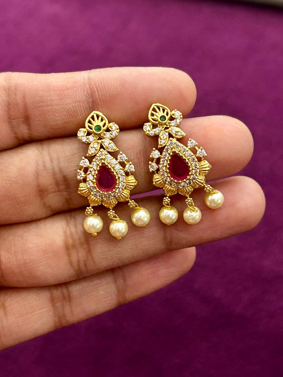 Gold earrings with red and green gemstones held between fingers against a purple background