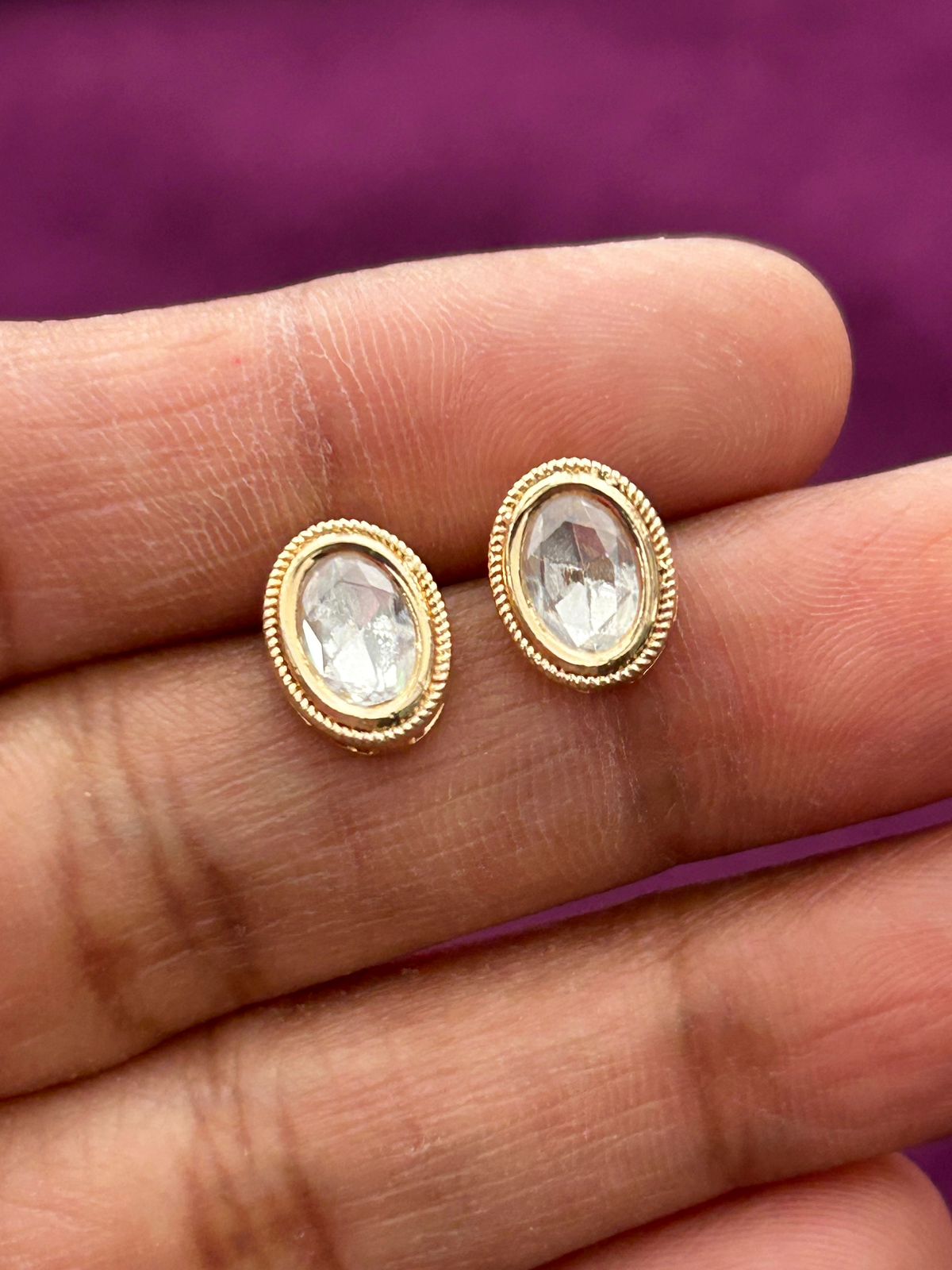 Gold earrings with clear gemstones held between fingers against a purple background