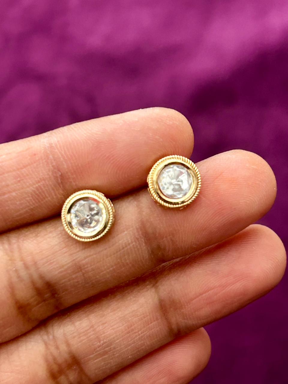 Gold earrings with clear stones held between fingers against a purple background