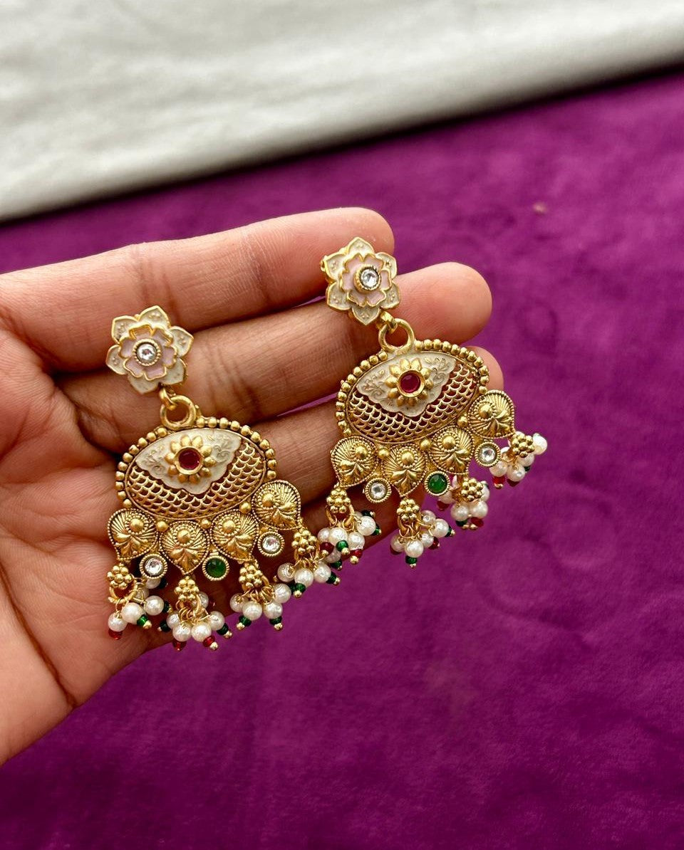 Gold earrings with floral designs held in a hand against a purple background