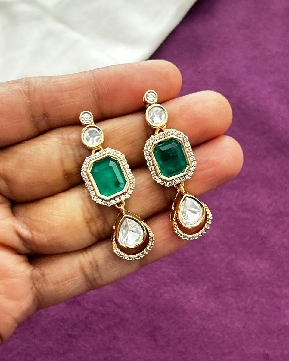 Gold earrings with green and clear gemstones held in a hand against a purple background