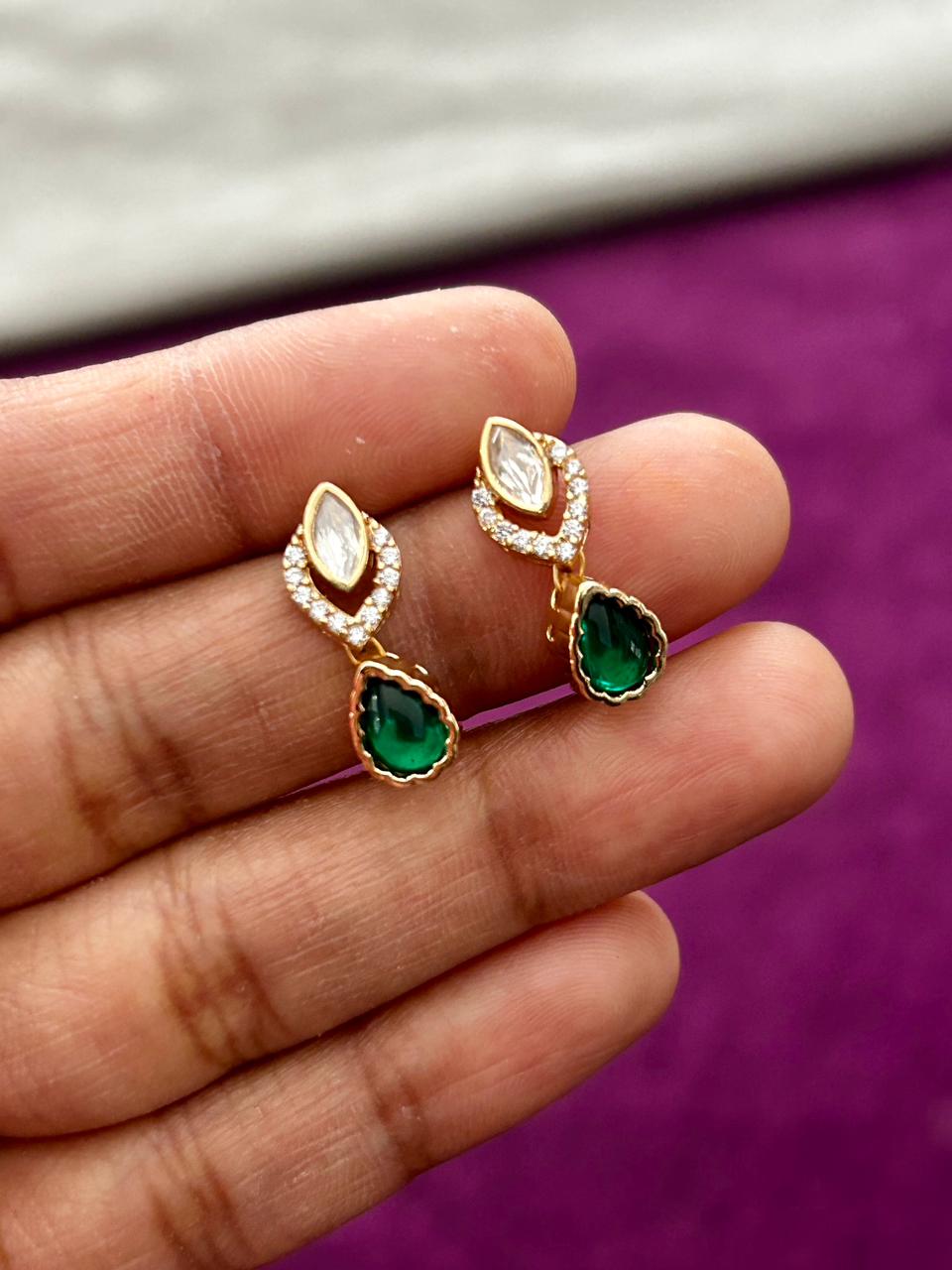 Gold earrings with green gemstones held between fingers against a purple background