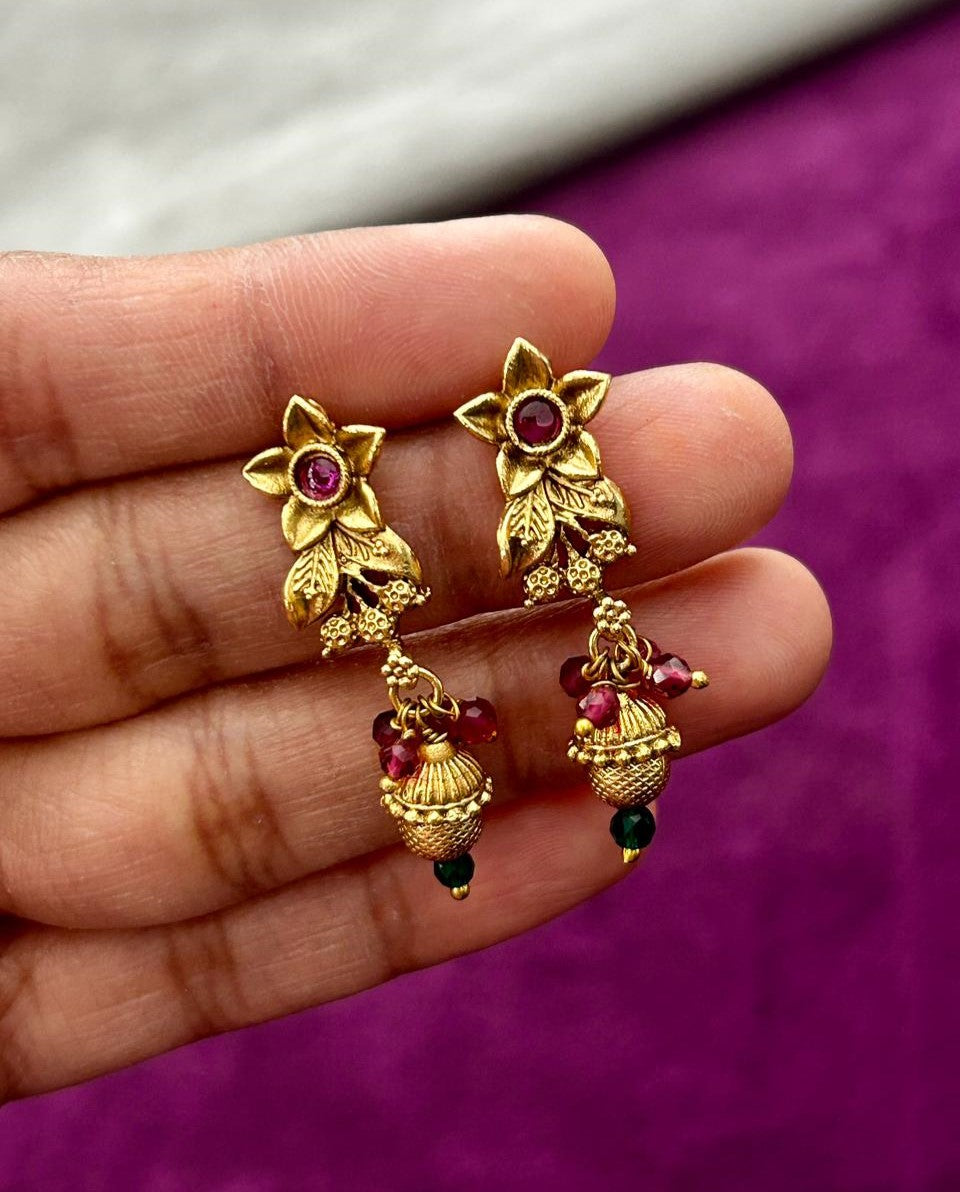Gold earrings with red and green gemstones held between fingers against a purple background