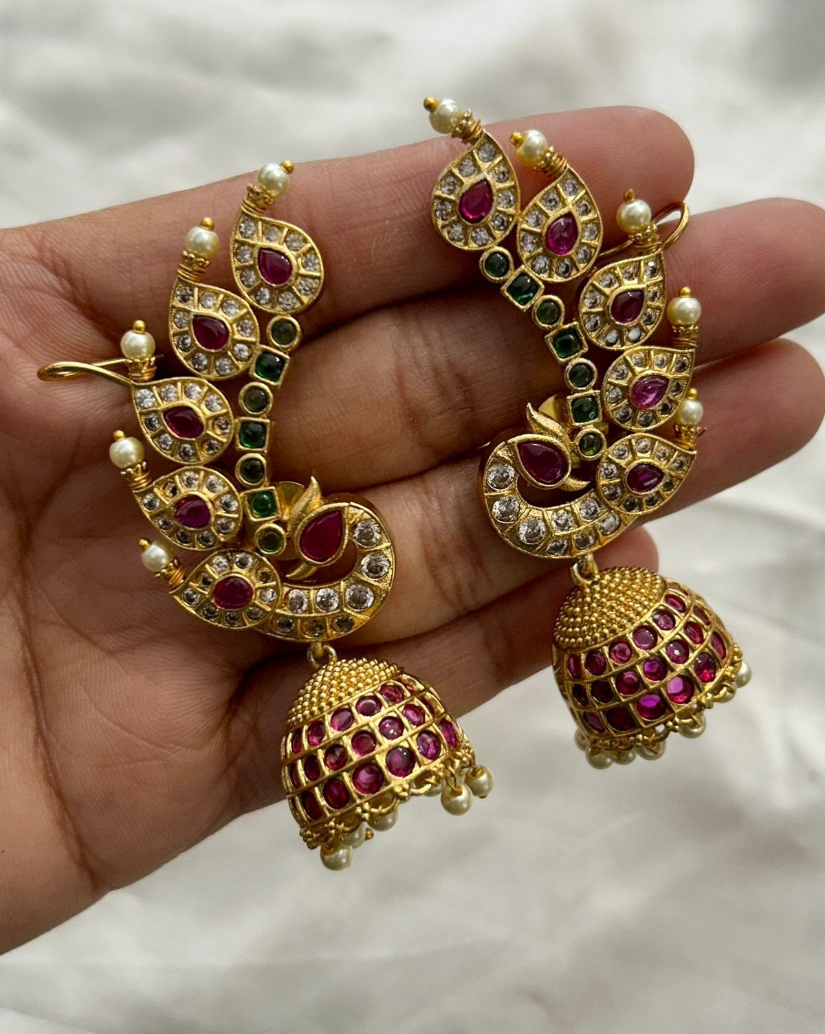Gold earrings with red and green gemstones held in a hand against a neutral background